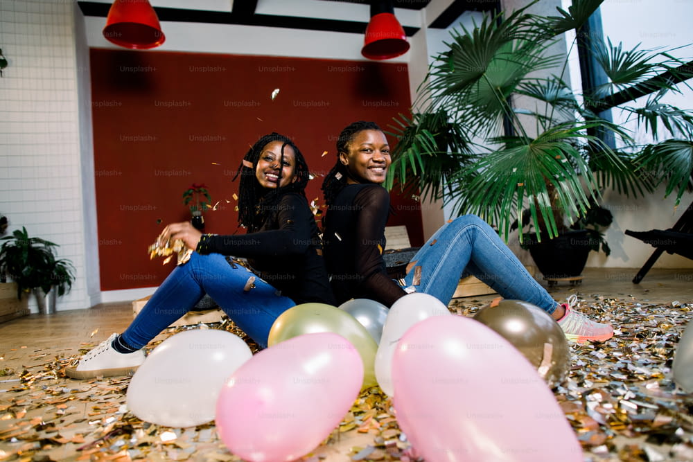 Zwei afrikanische Mädchen, fröhliche, stilvolle Freunde, die Silvester oder Geburtstagsfeier feiern, lehnen sich zurück und werfen ein Konfetti. Mode Eleganz Frauen genießen Zeit zu zweit.