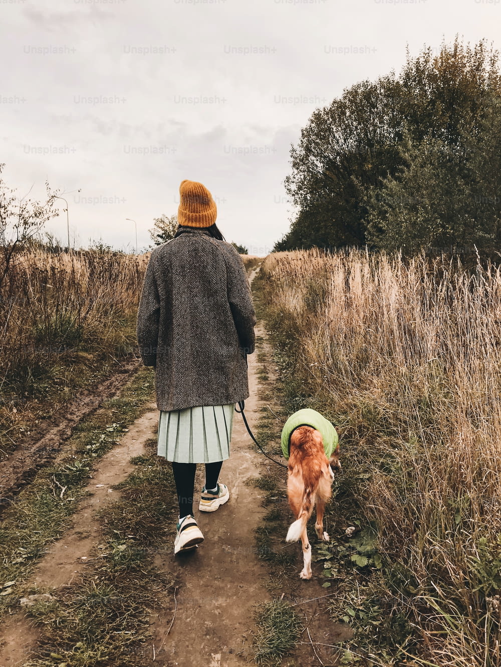 Stylish Hipster girl in yellow hat and coat walking with her golden dog in coat in autumn field among herbs. Woman in modern clothes walking with friend dog in countryside