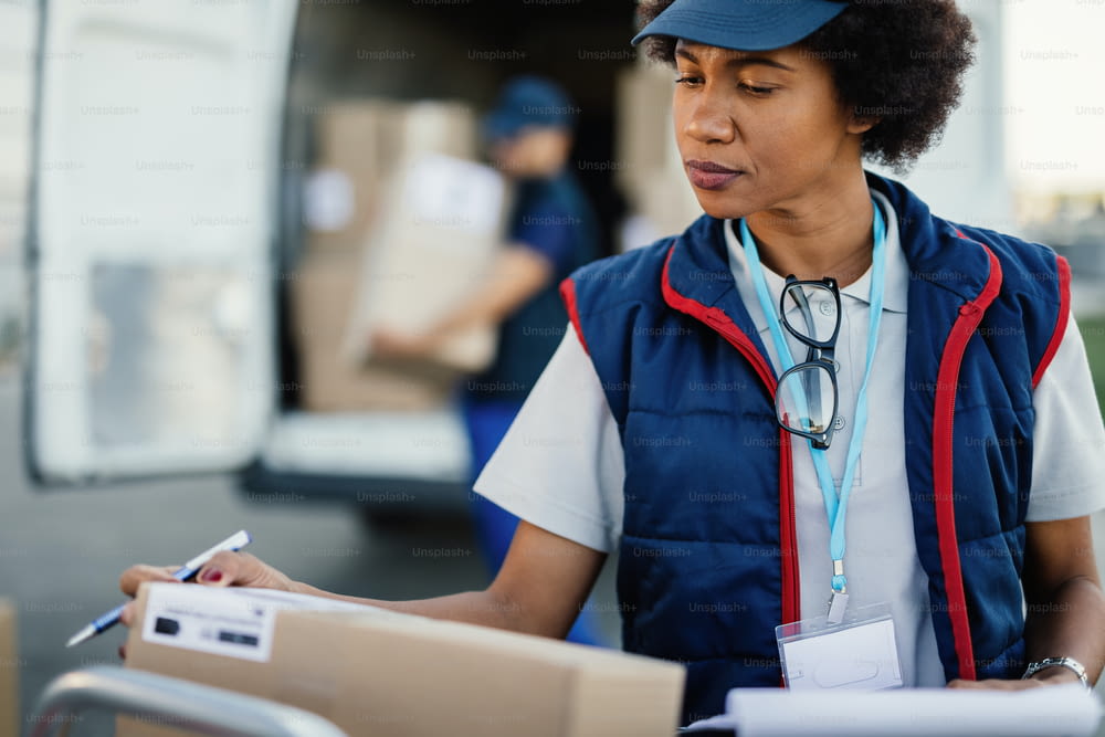 African American worker organizing delivering schedule and checking boxes before the delivery. Her colleague is loading packages in a van in the background.