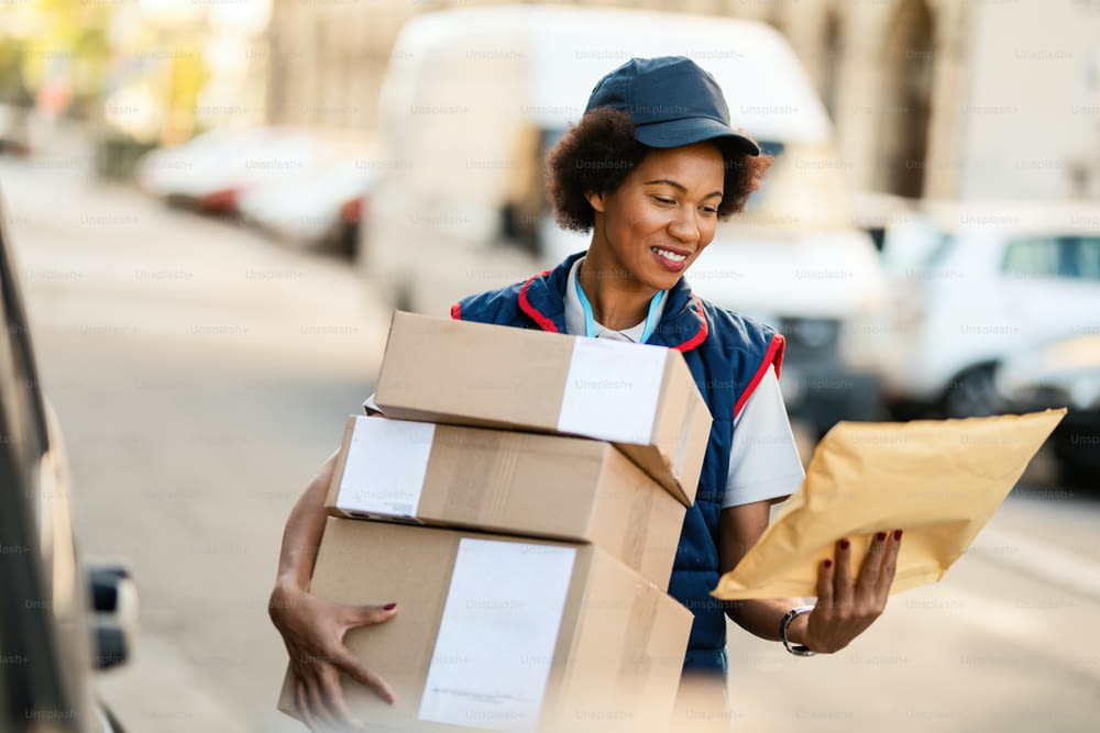 Happy African American delivery woman reading address on a package while making delivery in the city.