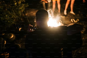 Hipster-Mann spielt akustische Gitarre und singt Lieder am großen Lagerfeuer im Camp, chillt mit Freunden im Wald. Gruppe von Menschen, die sich abends am Feuer entspannen, Camping in der Nähe des Sees