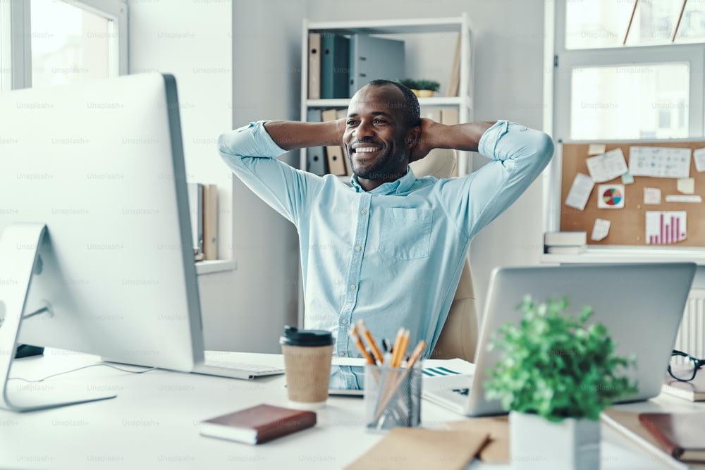 Bel giovane africano in camicia che tiene le mani dietro la testa e sorride mentre lavora in ufficio