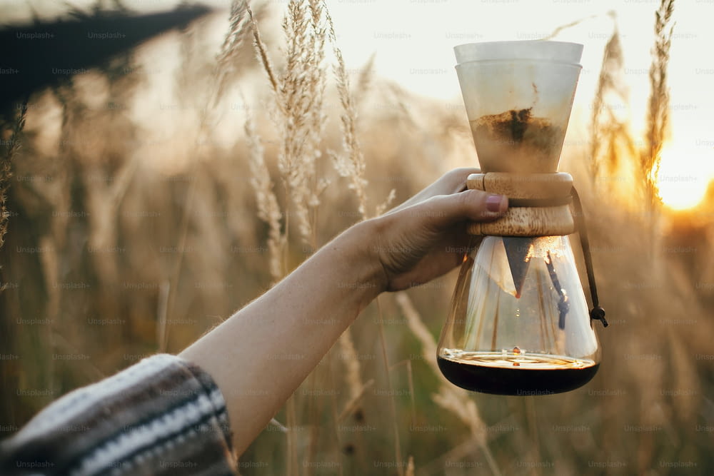 Alternative coffee brewing outdoors in travel. Hand holding hot coffee in glass flask with filter on background of sunny warm light in rural herbs. Atmospheric rustic tranquil moment. Copy space