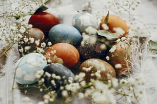 Stylish easter eggs in rustic nest on table. Natural dyed colorful easter eggs with spring white flowers and feathers on rural textile background. Happy Easter