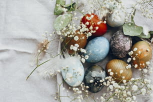 Stylish easter eggs in rustic nest on table flat lay. Natural dyed colorful easter eggs with spring white flowers and feathers on rural textile background. Happy Easter