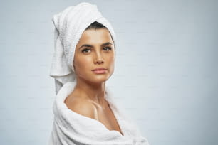 Seitenansicht einer hübschen Frau mit Handtuch auf dem Kopf und im Bademantel. Porträt einer brünetten Frau mit nackter Schulter in die Kamera, isoliert auf grauem Hintergrund. Beauty, Hautpflegekonzept.