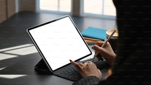 Cropped shot of business woman typing on keyboard of tablet and holding electric pen in hand while sitting at the modern working table with the comfortable office room as background.