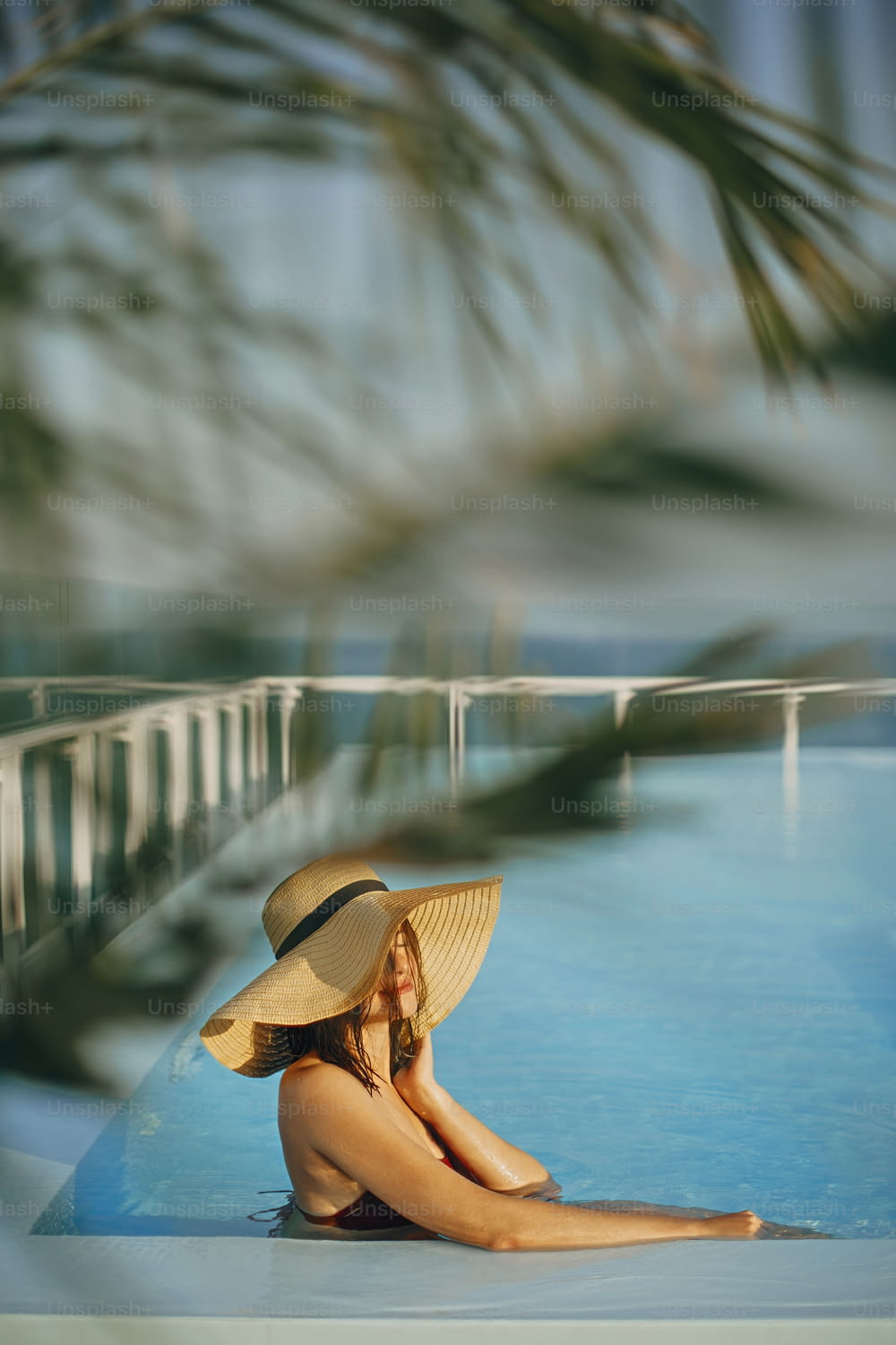 Stylish young woman in hat relaxing in pool under palm leaves and enjoying warm sunny light on summer vacation. Girl in sunhat on vacation in luxury tropical resort, swimming in pool on rooftop