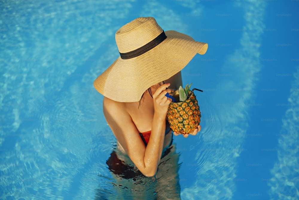 Bella giovane donna in cappello che beve un delizioso cocktail dall'ananas e si rilassa in piscina, vacanze estive. Ragazza che gode di sole caldo, nuoto in piscina sul tetto in un resort tropicale di lusso