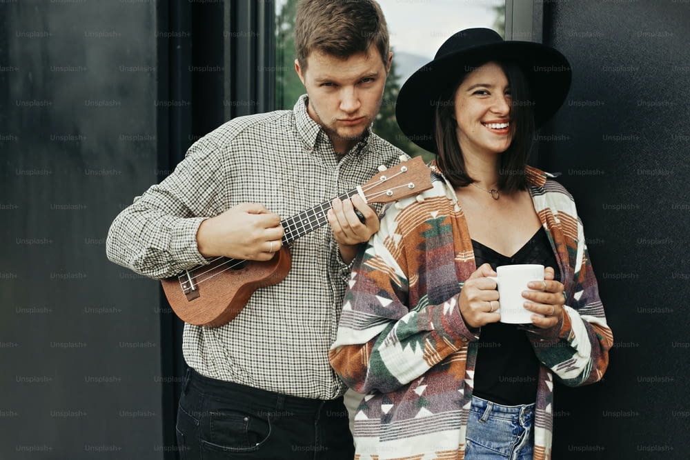 Stylish hipster couple with morning coffee and ukulele smiling, relaxing on background of modern cabin with big windows in mountains. Happy young family enjoying vacation in woods