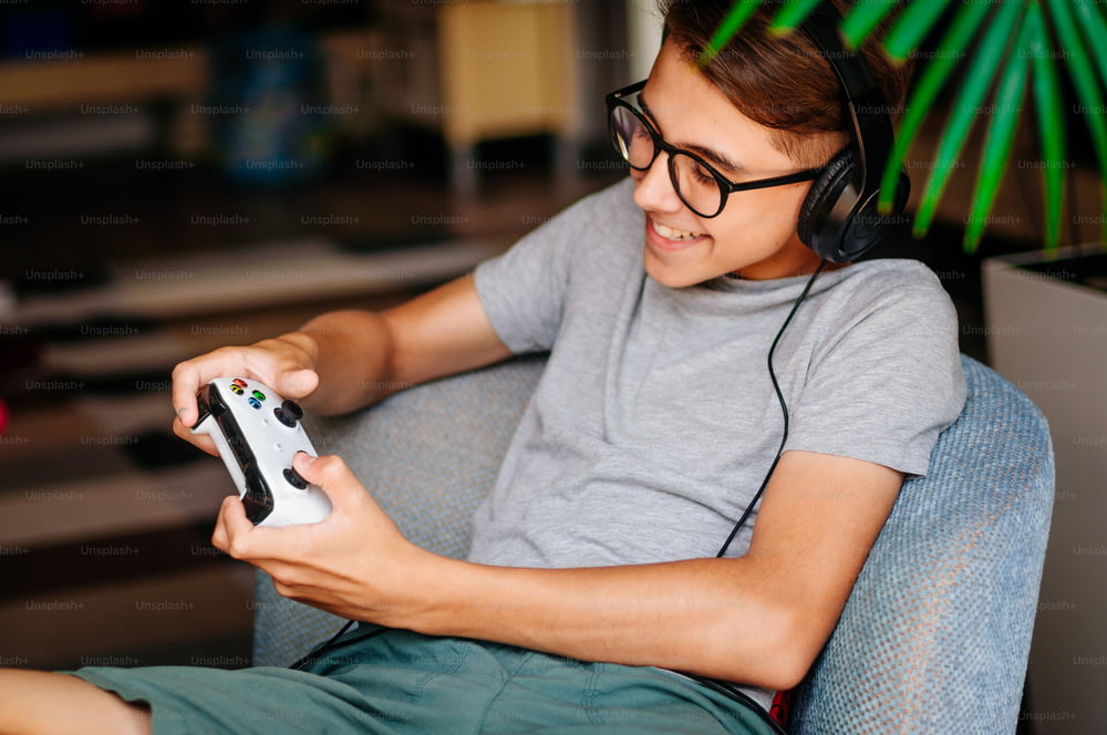 Menino adolescente sorridente que joga o console com fones de ouvido sentados na cadeira interna