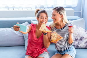 Amigos comiendo pizza y sonriendo para una selfie. Están compartiendo pizza y haciendo fotos selfie en un teléfono inteligente móvil. Están de fiesta en casa, comiendo pizza y divirtiéndose.