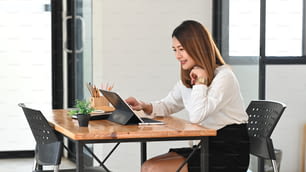 Junge schöne Frau, die als Sekretärin mit einem Computer-Tablet mit Tastaturgehäuse arbeitet, während sie am hölzernen Arbeitstisch mit geordnetem Büro als Hintergrund sitzt.