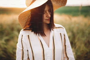 Retrato de una mujer hermosa con sombrero que disfruta de la luz dorada de la puesta del sol en el prado de verano. Muchacha rústica con estilo en vestido de lino que se relaja por la noche en el campo. Vida rural lenta. Momento atmosférico