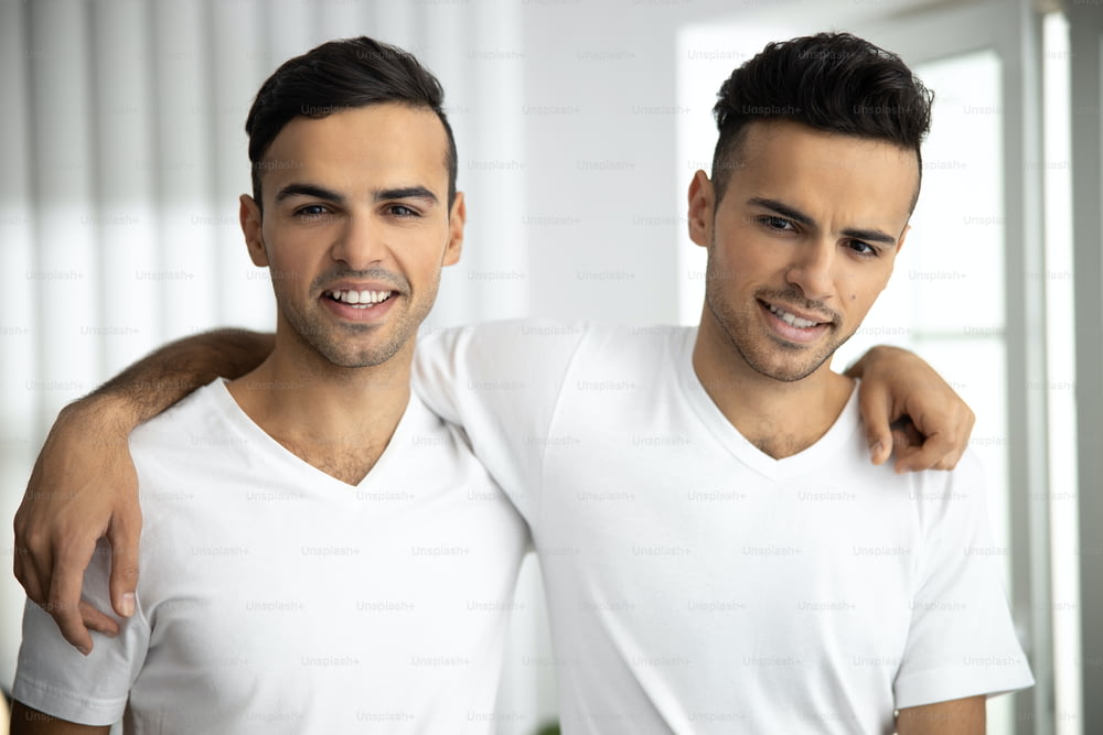 Cintura para cima de gêmeos masculinos felizes e amigáveis em camisetas brancas abraçando em casa stock foto