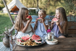 Three cute happy smiling girls, sisters, fiends, sitting at the table on vintage wooden bench and eating watermelon outdoors, in tent, stylish boho wigwam on background. Countryside summer holidays.