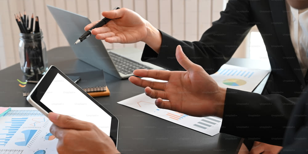 Cropped image of business people working together with computer tablet, laptop and graphic charts while sitting together at black meeting table over modern office as background.