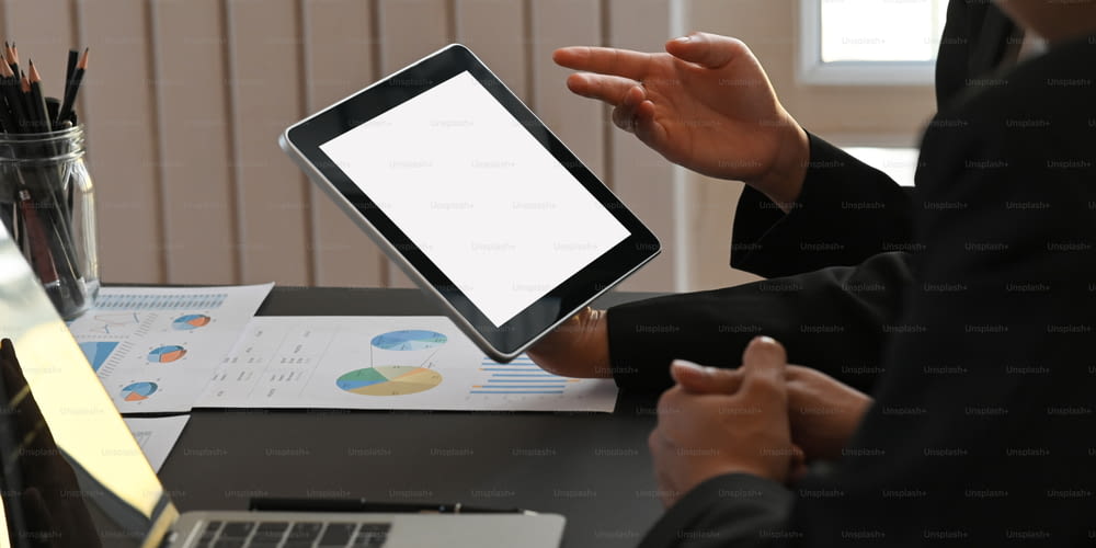 Cropped image of business people working together with computer tablet, laptop and graphic charts while sitting together at black meeting table over modern office as background.