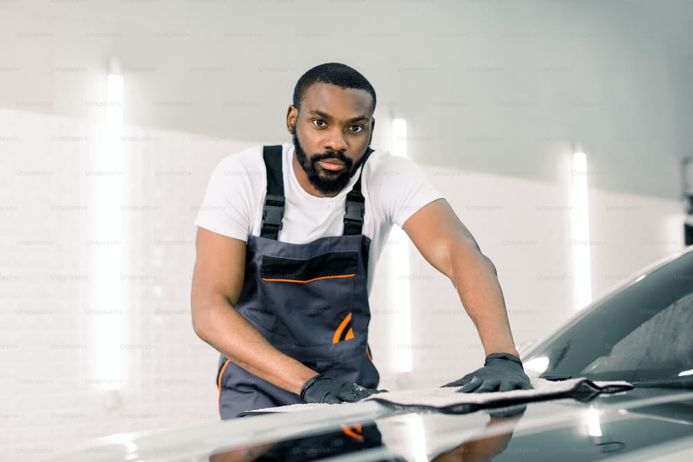 Car detailing and cleaning concept. Good-looking African male car wash worker, wearing special overalls and gloves, wipes and polishes the car hood with microfiber cloth, looking at camera.