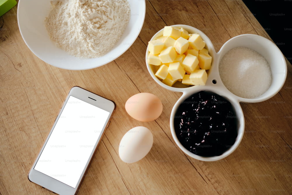 テーブルの上のケーキの材料は、白い画面のスマートフォンでレシピに従っている間、調理する準備ができています。バター、宝石、卵、床