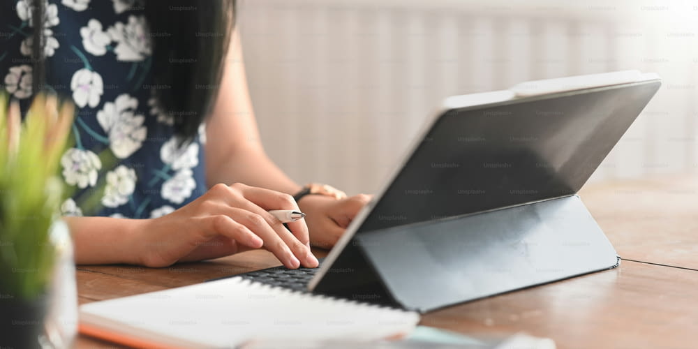 Cropped image of stylish woman holding a stylus pen and typing on computer tablet keyboard case while sitting at the wooden working desk over comfortable room as background.
