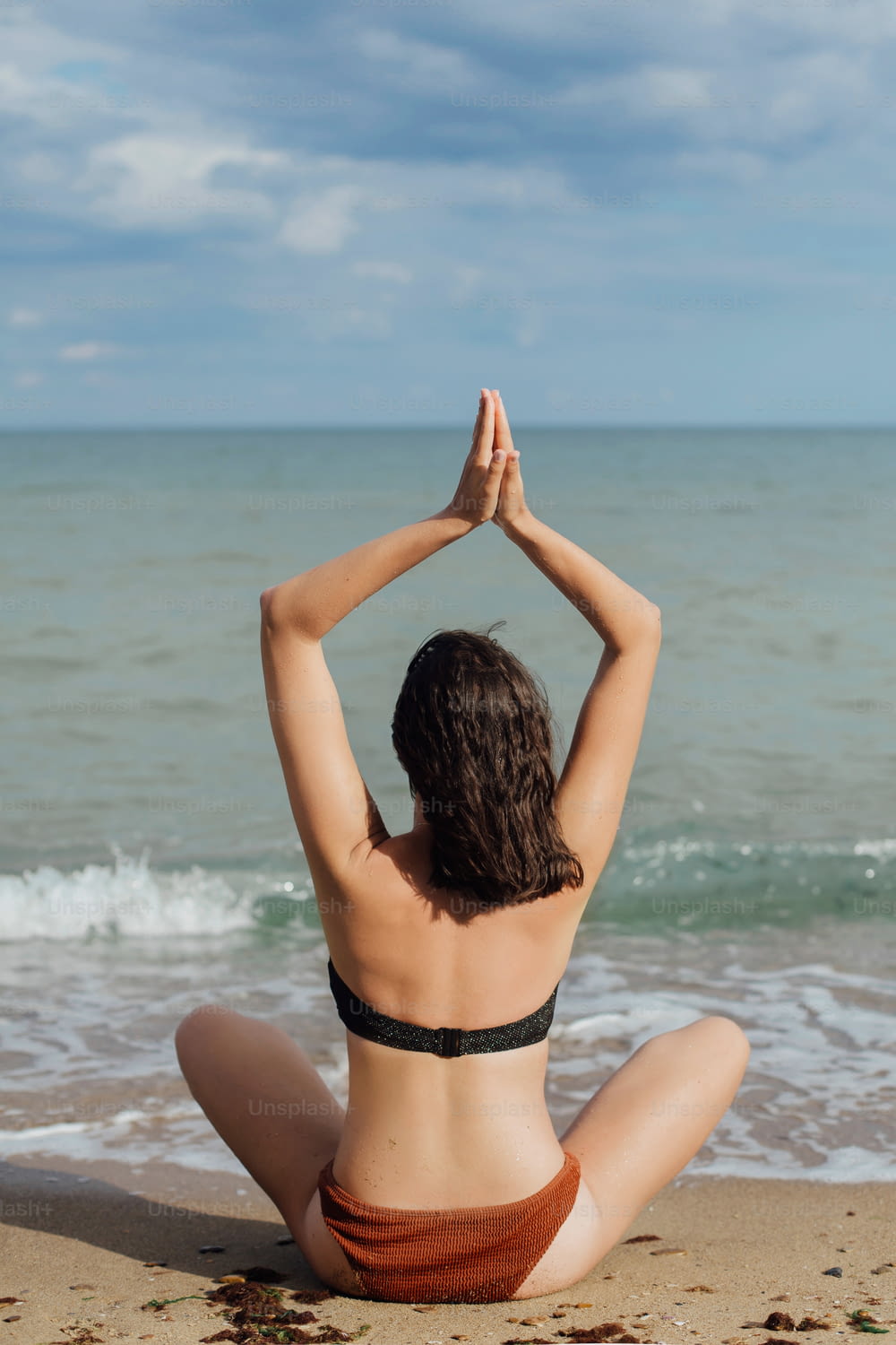 Young fit woman practicing yoga on the beach, sitting on sand and looking at sea waves. Girl meditating on summer vacation. Mental health and self care concept