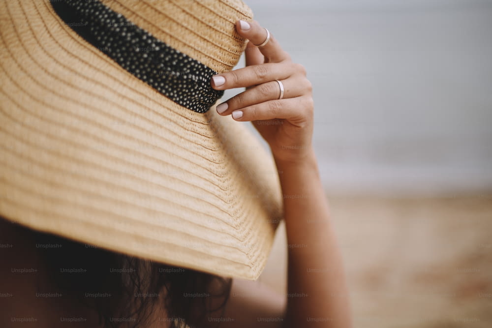 夏休みと旅行。ビーチに座って、帽子をクローズアップした手を持つ女の子。麦わら帽子をかぶったファッショナブルな若い女性が海の近くの砂浜でリラックスしています。のんびり。テキスト用のスペース