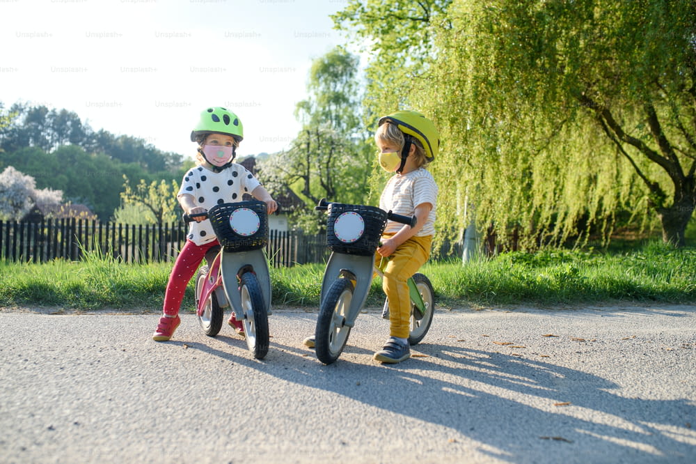 Niños pequeños niño y niña con máscaras faciales jugando al aire libre con bicicleta, concepto de coronavirus.