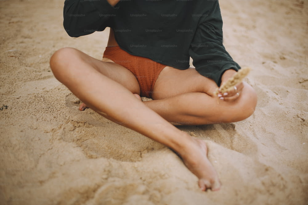 Hipster-Mädchen, das Kräuter hält und am Strand sitzt, abgeschnittener Blick auf gebräunten Beinen. Stilvolle Boho-Frau in modernem Badeanzug und Pullover, die sich am Meer entspannt. Sommerferien. Achtsamkeit