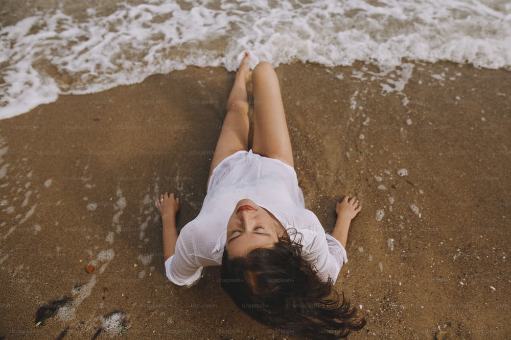 젖은 흰 셔츠를 입은 젊은 여자가 튀는 파도에 해변에 누워 있다. 평면도. 해변에서 휴식을 취하고 파도를 즐기는 세련된 검게 그을린 소녀. 여름 방학. 마음 챙김과 평온