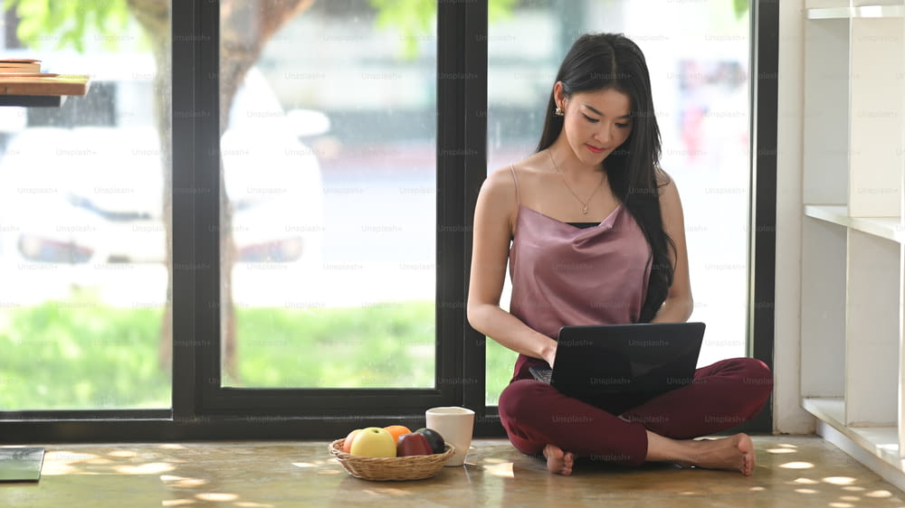 Asian woman using her laptop and sitting on floor in living room.