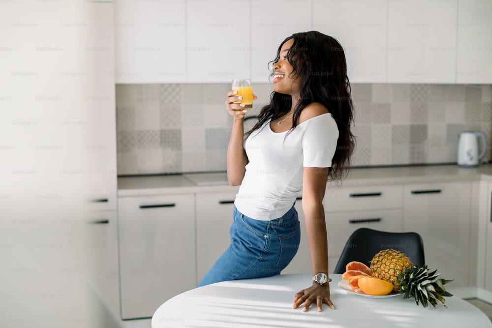 Giovane bella donna afroamericana che tiene il vetro con un succo d'arancia, guardando la finestra, in posa alla cucina moderna leggera a casa. Concetto di stile di vita sano, colazione biologica naturale in cucina.