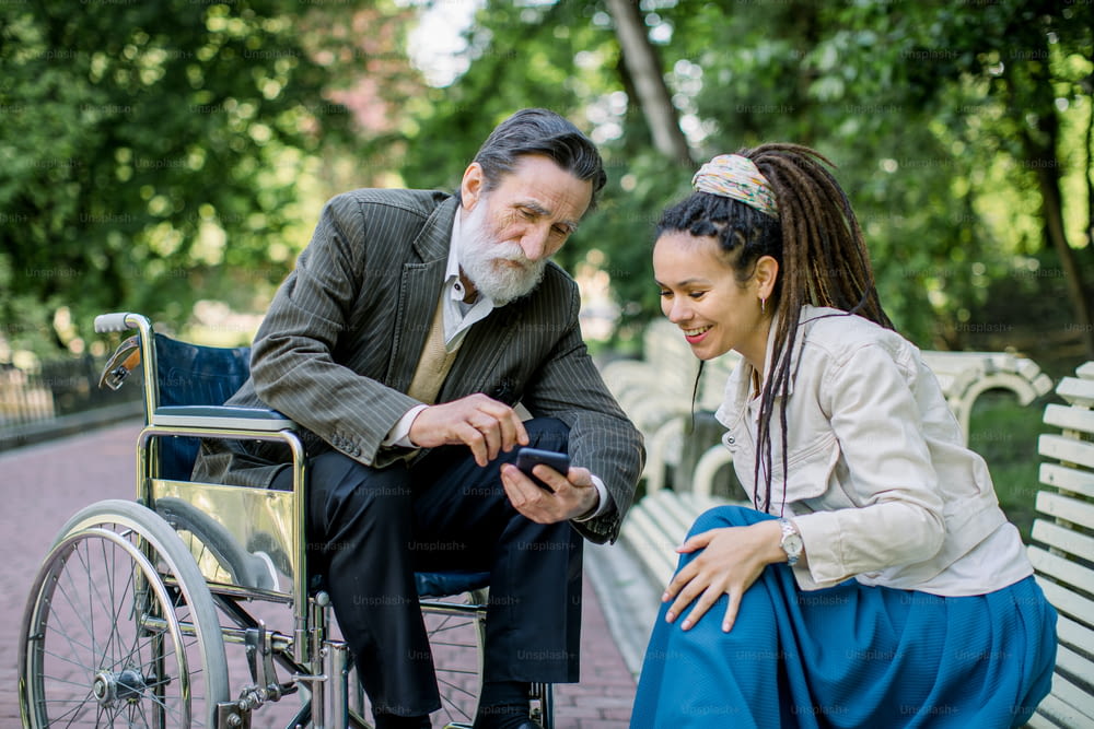 Portrait of smiling young hipster woman with long dreadlocks, sitting on the bench in park, enjoying time with her disabled grandfather on wheelchair, showing photos on the smart phone.