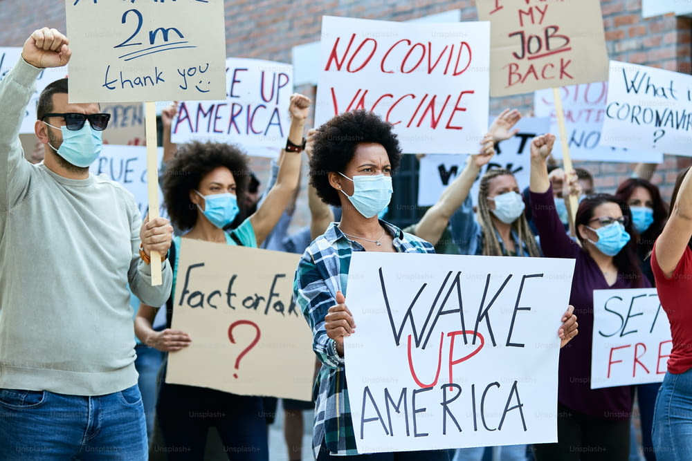 코로나 바이러스 전염병 기간 동안 도시 거리에서 시위하는 안면 마스크를 쓴 사람들의 불쾌한 군중. 초점은 Wake up America라는 문구가 새겨진 플래카드를 들고 있는 흑인 여성에게 맞춰져 있습니다.