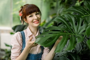 背景に植物と温室に立っている間、彼女の顔の近くにモンステラの葉を保持してカメラにポーズをとって、笑顔の魅力的な赤毛の女性庭師の肖像画。