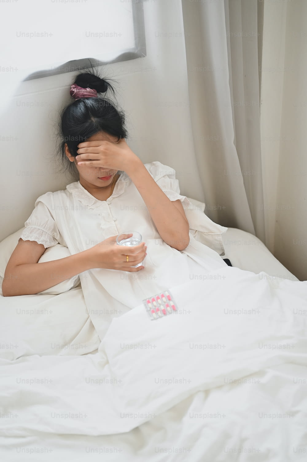 Eine kranke Frau hält ein Glas Wasser und konsumiert eine Pille, während sie auf dem Bett im Schlafzimmer liegt.