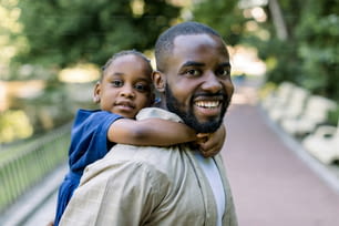 Smiling handsome African American father carrying his cute little daughter on back outdoors in city park. Daddy carrying her daughter on piggyback in the nature
