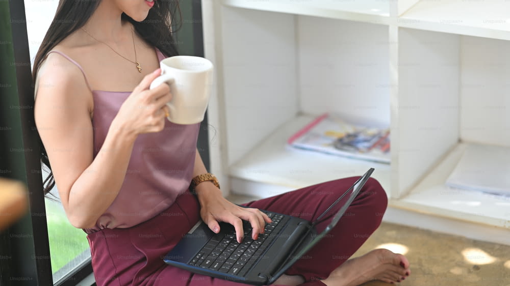 Cropped image woman is drinking coffee and using a computer laptop while sitting in the living room.