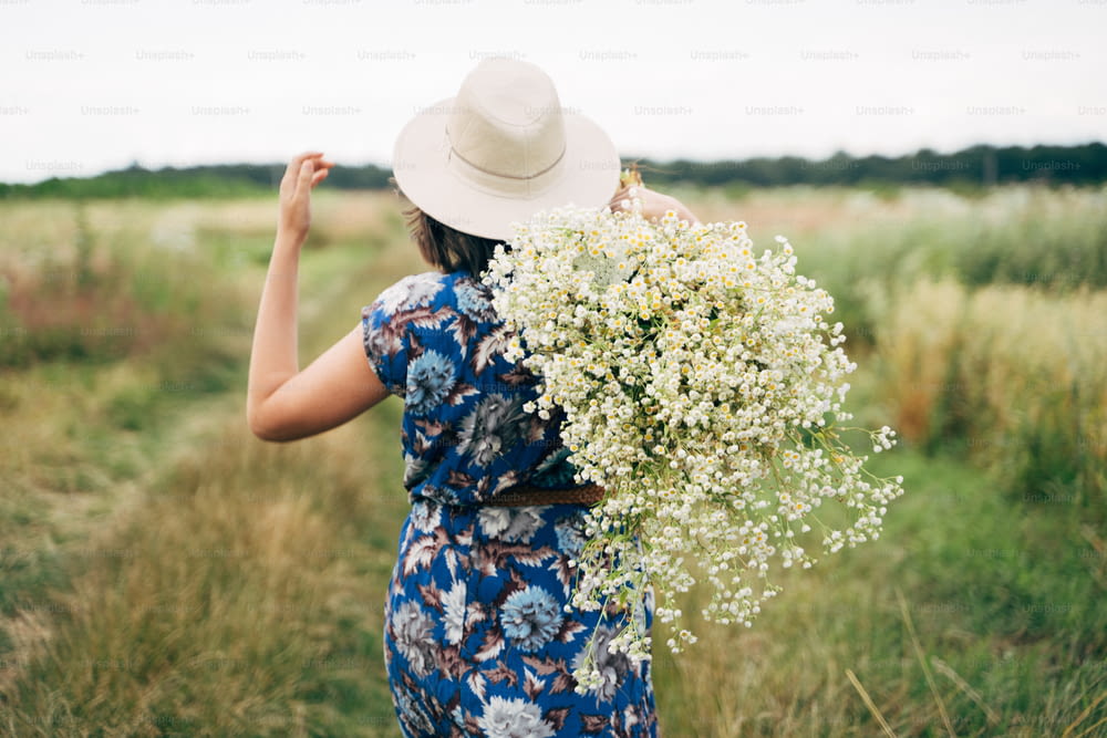 Verano en el campo. Mujer joven con estilo en vestido vintage azul y sombrero caminando con flores silvestres blancas en el prado de verano. Hermosa muchacha con un gran ramo de margaritas en el campo, vista posterior.