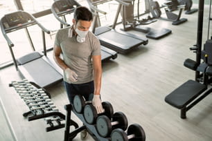 Männlicher Athlet trägt Gesichtsmaske und desinfiziert Handgewichte in einem Fitnessstudio während der COVID-19-Epidemie.