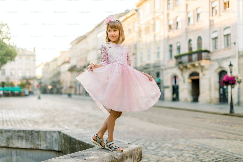 Retrato de cuerpo entero de moda de una linda niña rubia, con vestido rosa y flor en el cabello, posando a la cámara, mientras está de pie en una antigua fuente de piedra en la antigua ciudad europea.