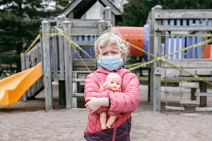 Menina caucasiana triste em máscara facial com brinquedo de bebê no playground fechado ao ar livre. Área de recreação infantil trancada com fita amarela de advertência em Toronto, Canadá. Quarentena de distanciamento social do coronavírus.