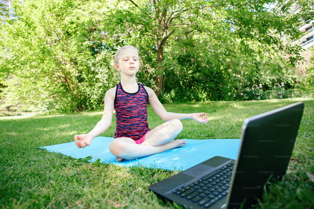 Girl child practicing yoga outdoor online. Video sport workout on Internet. Kid learning fitness training with laptop. New normal. Social distance and education at coronavirus. Stress relief concept.