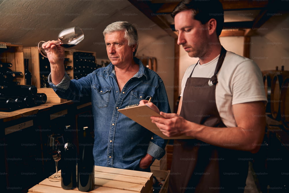 Calm Caucasian man standing in a winery cellar with his younger colleague and looking at the wine while tilting a glass