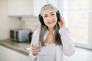 Portrait of happy smiling mature granny with long gray hair, standing in modern light kitchen with glass of water, listening to the music or audio book using headphones