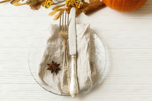 Stylish and zero waste Thanksgiving table setting. Plate with cutlery, linen napkin, anise and autumn leaves, pumpkin, autumnal flowers on white table. Rustic autumn wedding table set.