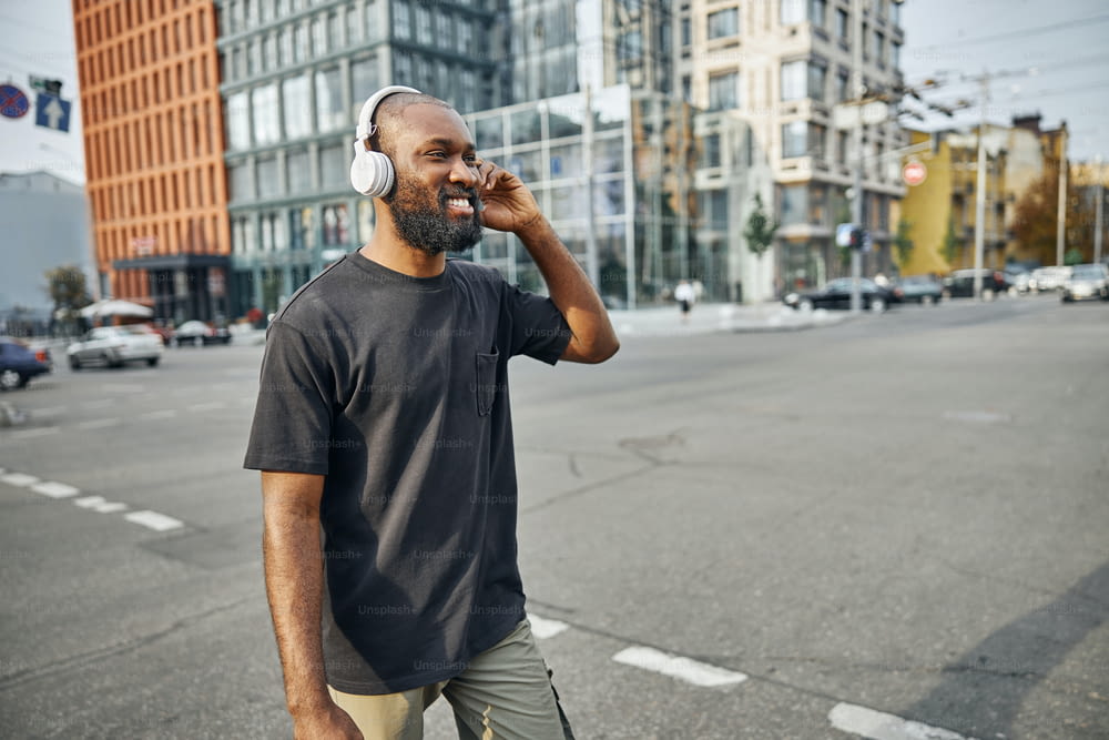 Zufriedener junger Mann in Freizeitkleidung, der in der Innenstadt spaziert und die drahtlosen Kopfhörer auf seinem Kopf berührt. Website-Banner