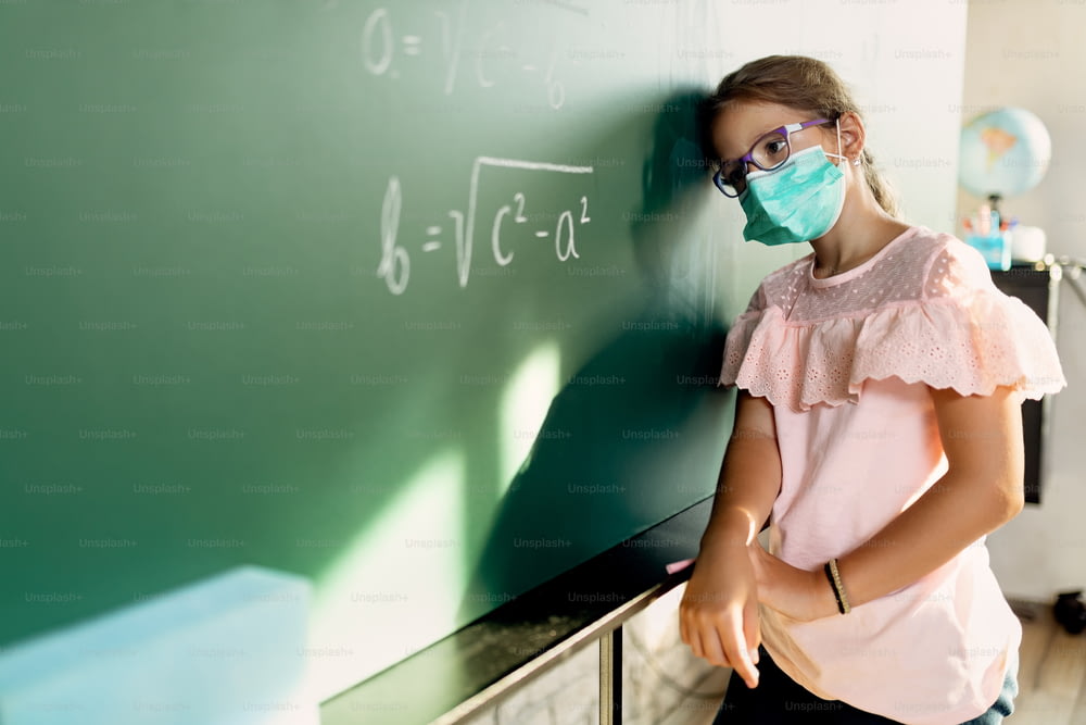 Schoolgirl with protective face mask feeling sad because she can't solve math assignment on blackboard.