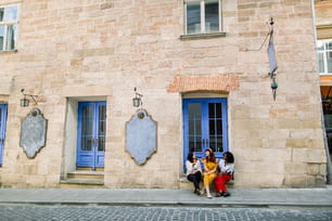 Three young multiethnic women tourists, Muslim, African and Caucasian, having fun on the street in old European city, sitting on the stairs under blue cafe door, and eating fresh croissants.