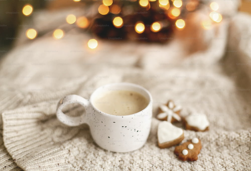 Warm coffee in stylish cup on background of cozy knitted sweater with christmas gingerbread cookies and warm lights. Atmospheric winter hygge. Happy Holidays